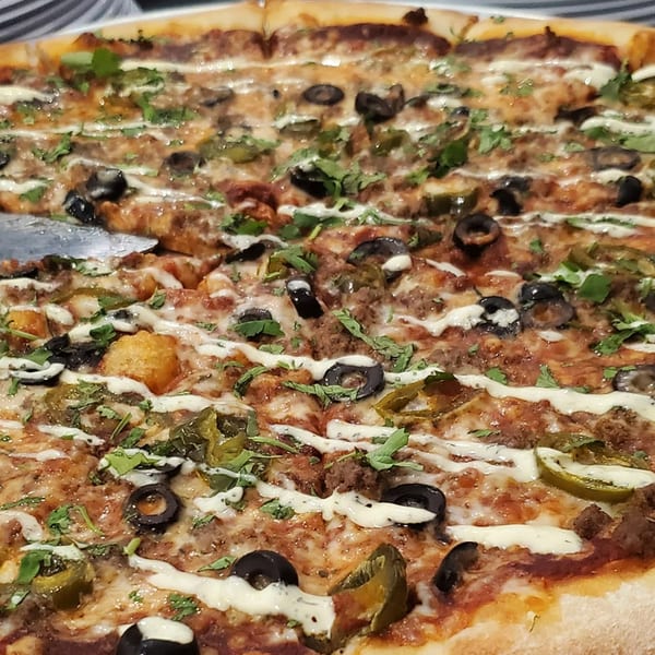 A close-up of a Slice On Oak pizza with ground meat, black olives, green peppers, and white sauce.