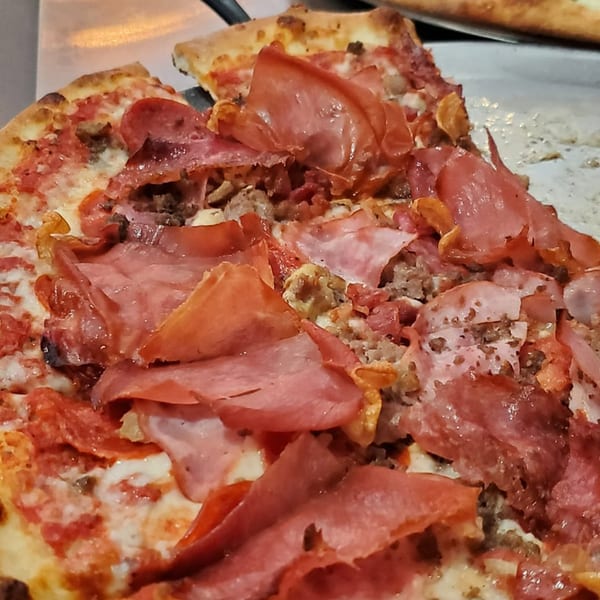 Close-up of a Slice On Oak pizza with pepperoni, sausage, and thinly sliced ham.